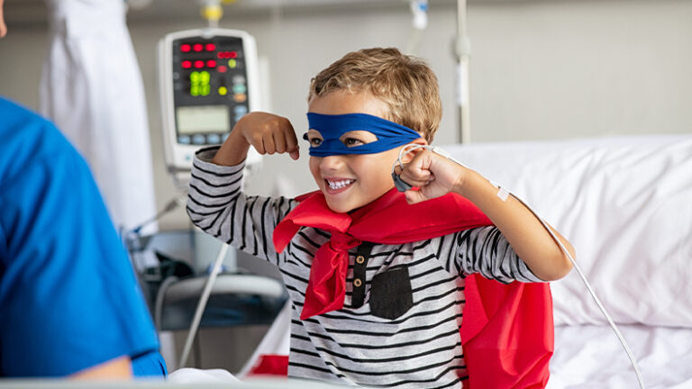 Cheerful strong little boy wearing blue eyeband and red cape like superhero sitting in hospital bed playing with nurse. Playful child gesturing dressed in superhero costume at clinic overcome adversity and health challenge. Happy smiling kid playing at hospital.